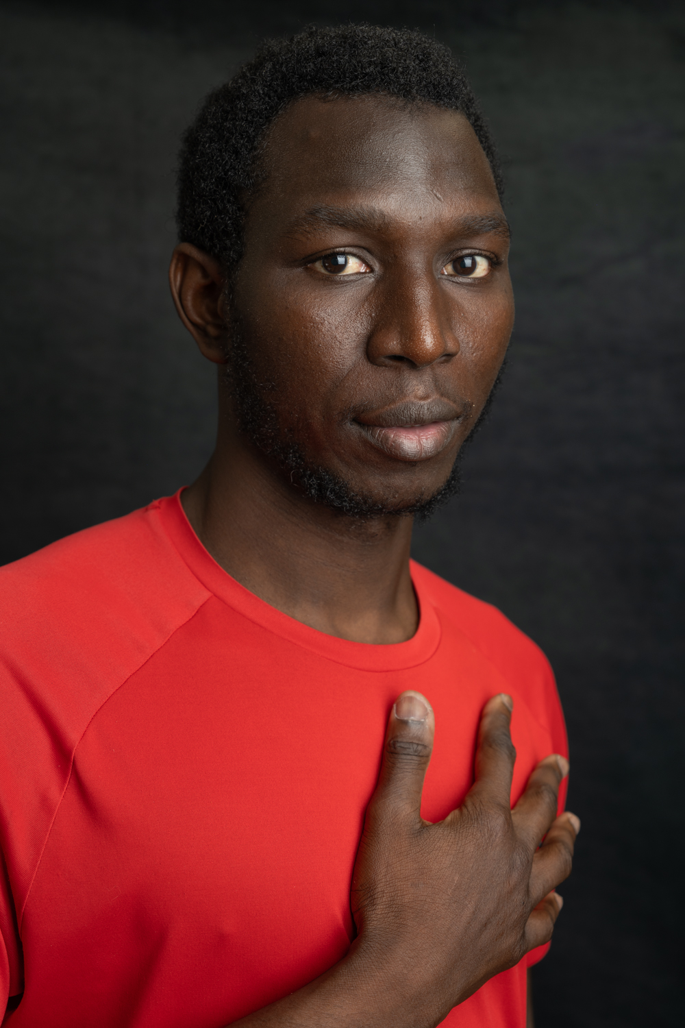 A photographic portrait of a man with short Afro hair. He has his hand resting on his chest and is wearing a bright red t-shirt.