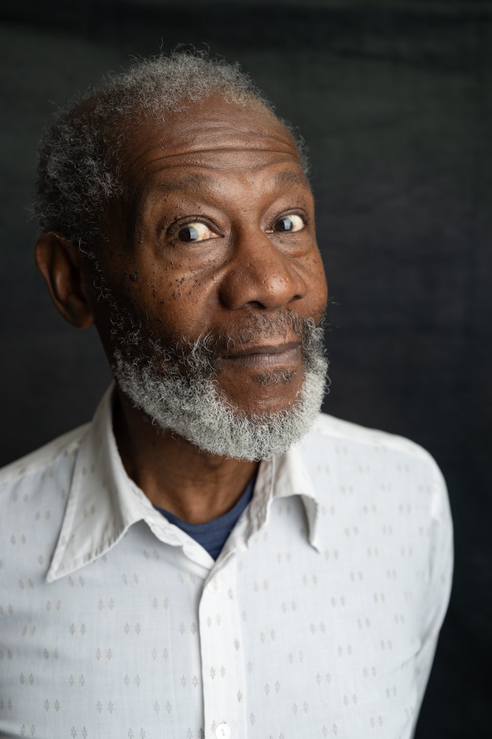 A photographic portrait of a man with white Afro hair and a white beard. He is wearing a white shirt and is leaning torward the camera with his eyes wide.