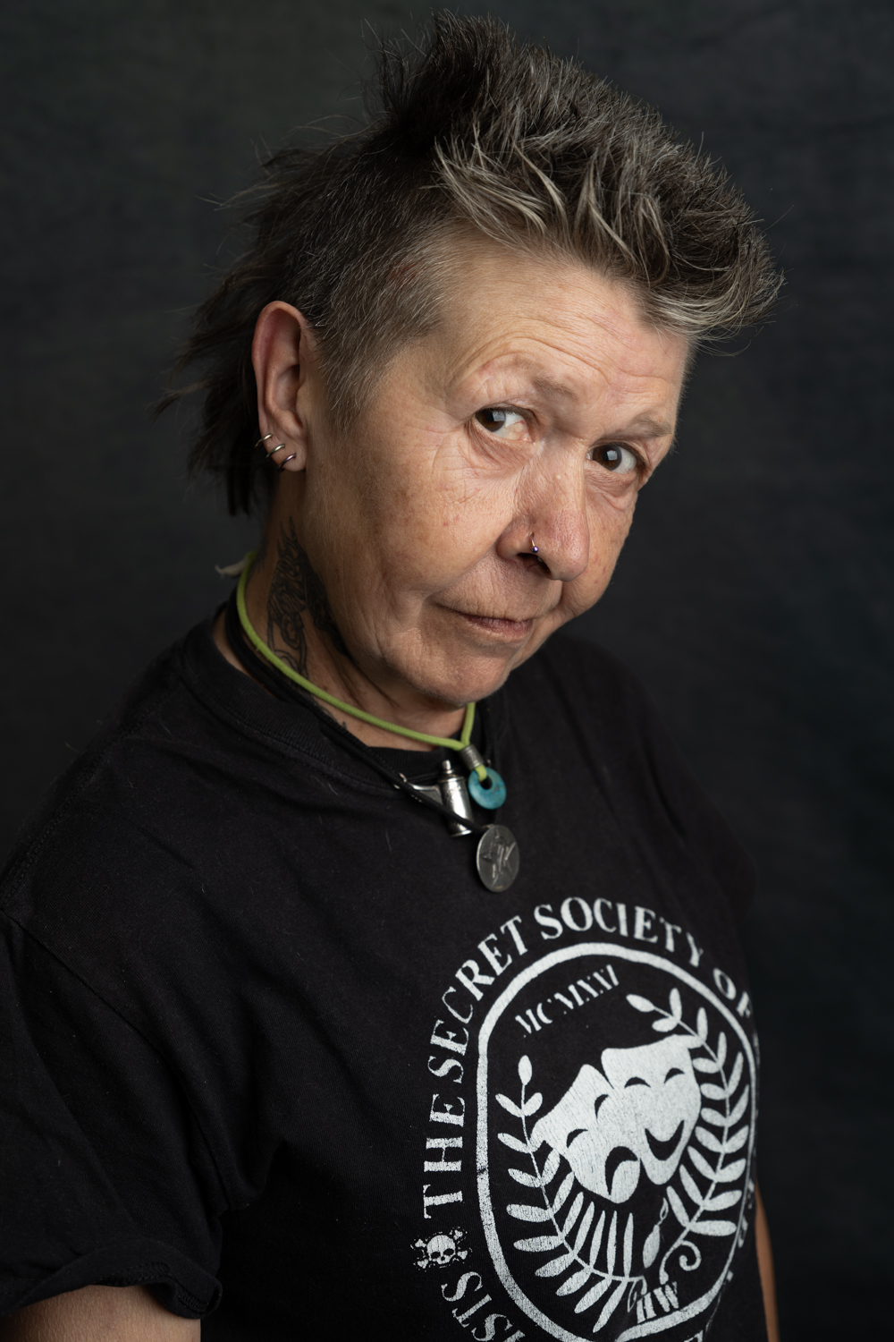 A photographic portrait of a woman with short grey hair. She is wearing a black t-shirt with a white design on the front, and lots of jewelry.