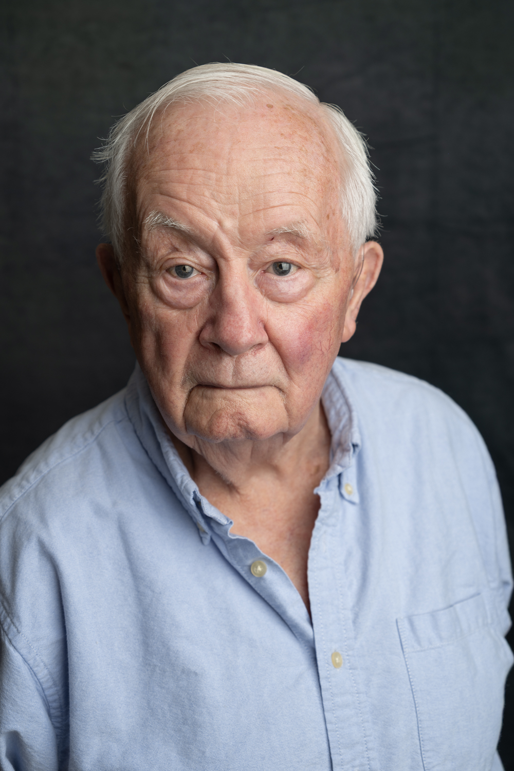 A photographic portrait of a man with short white hair. He is wearing a blue collared shirt and is not smiling.
