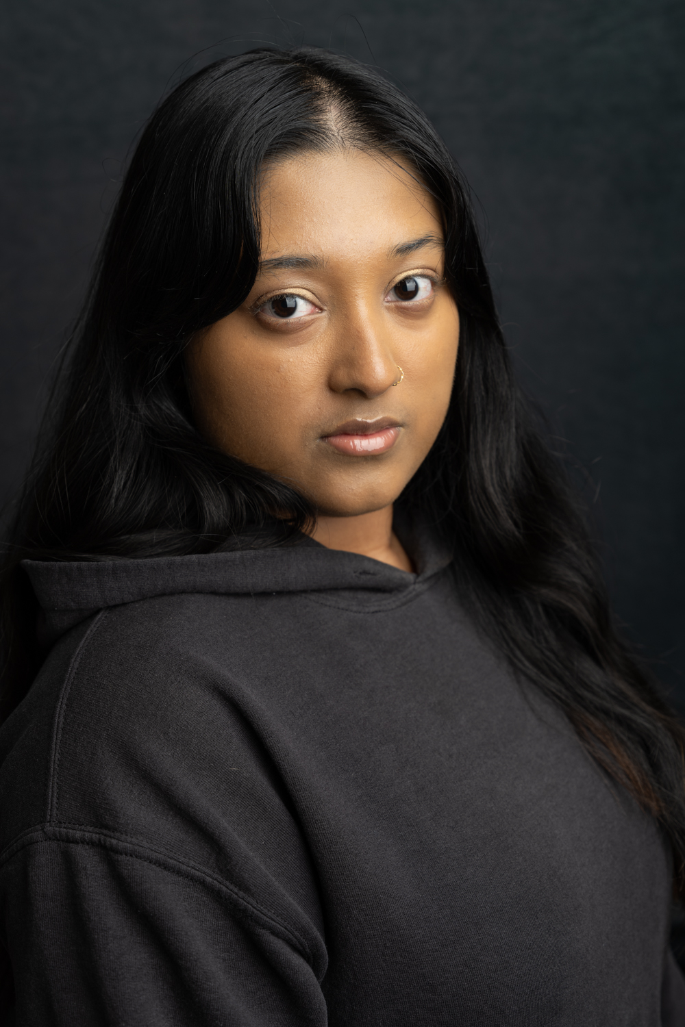 A photographic portrait of a woman of South Asian descent, with long black hair and a grey hoodie. She is not smiling, but is looking directly at the camera.