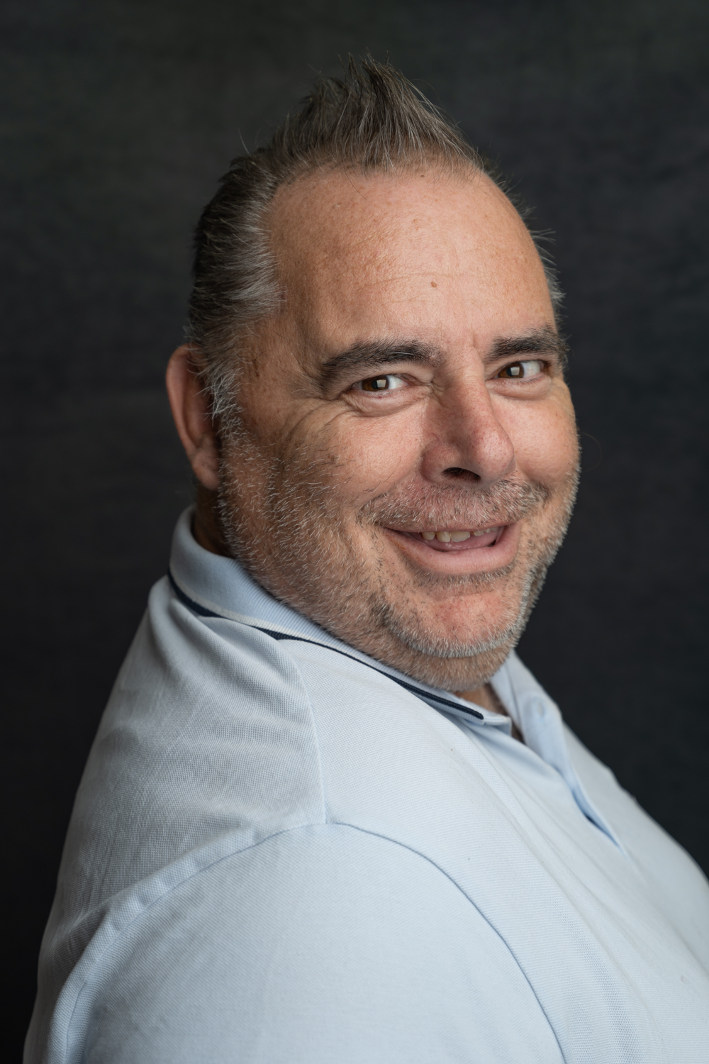A photographic portrait of a man with grey spiked hair and a big smile. He is wearing a light blue polo top and has grey stubble on his face.