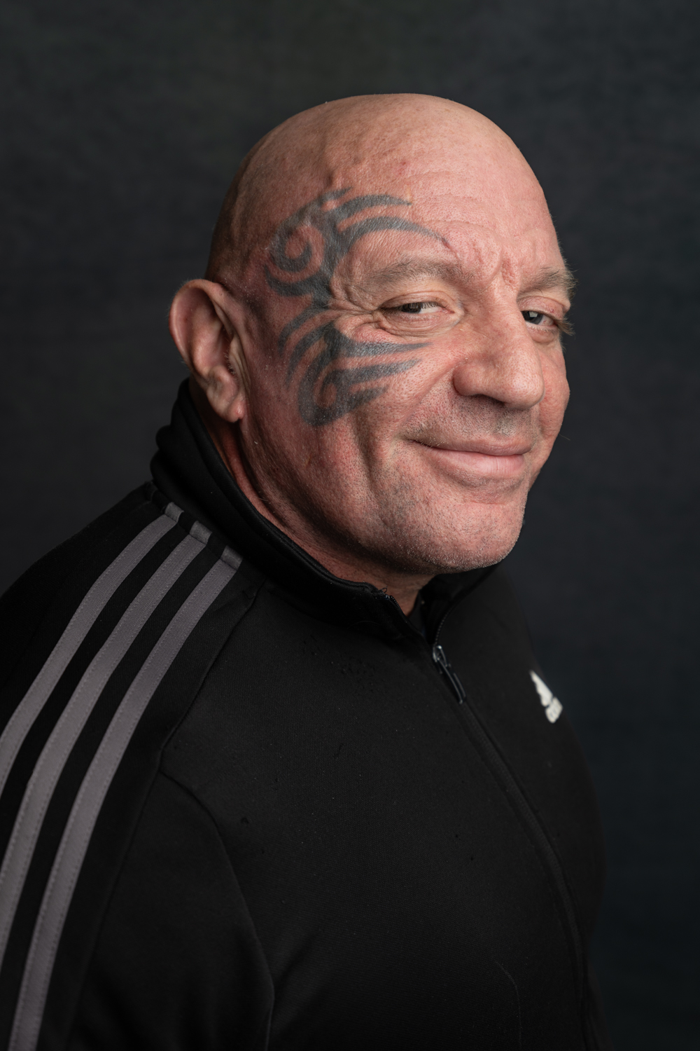 A photographic portrait of a bald man, wearing a black quarter zip jumper. He has a large tattoo around one of his eyes and is smiling at the camera.