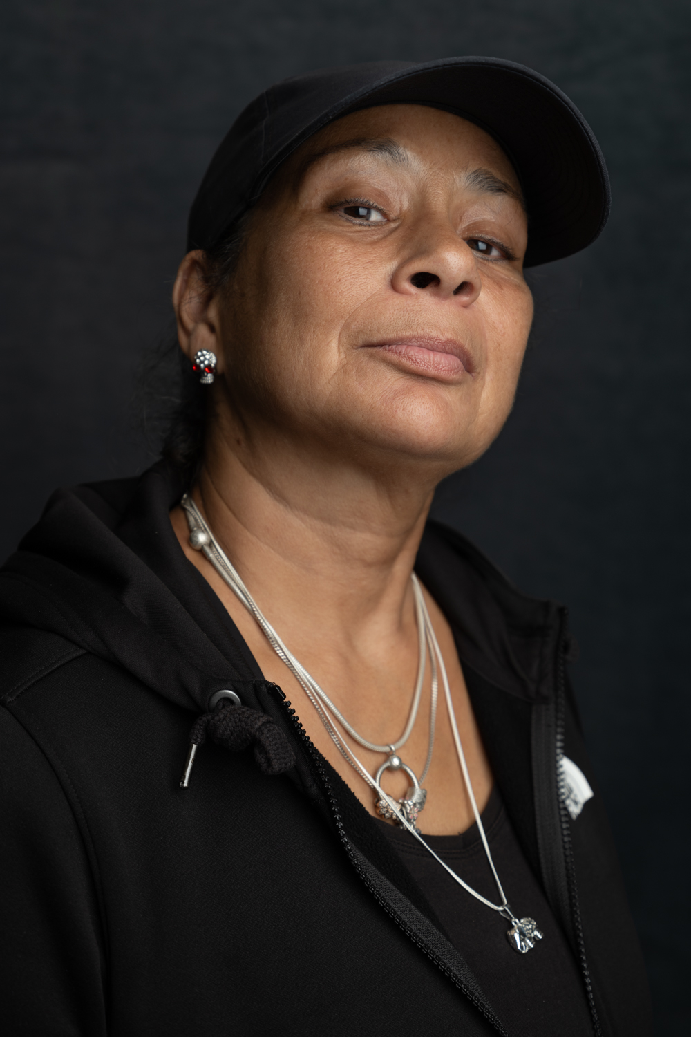 A photographic portrait of a woman wearing a black hoodie and a black baseball cap. She has her chin tilted slightly upwards and is wearing multiple necklaces and has a sparkly silver earring.
