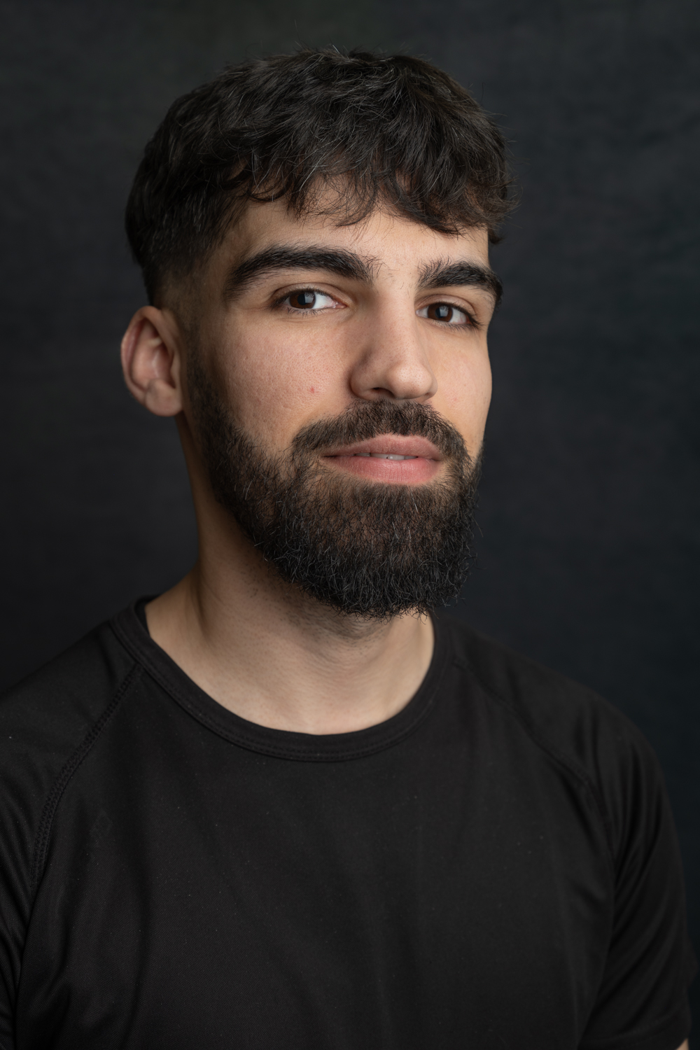 A photographic portrait of a man with dark hair and a thick dark beard. He is wearing a black t-shirt and is smiling slightly.