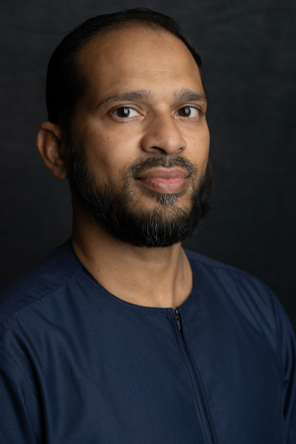 A photographic portrait of a man wearing a blue top. He has short dark hair and a short dark beard.