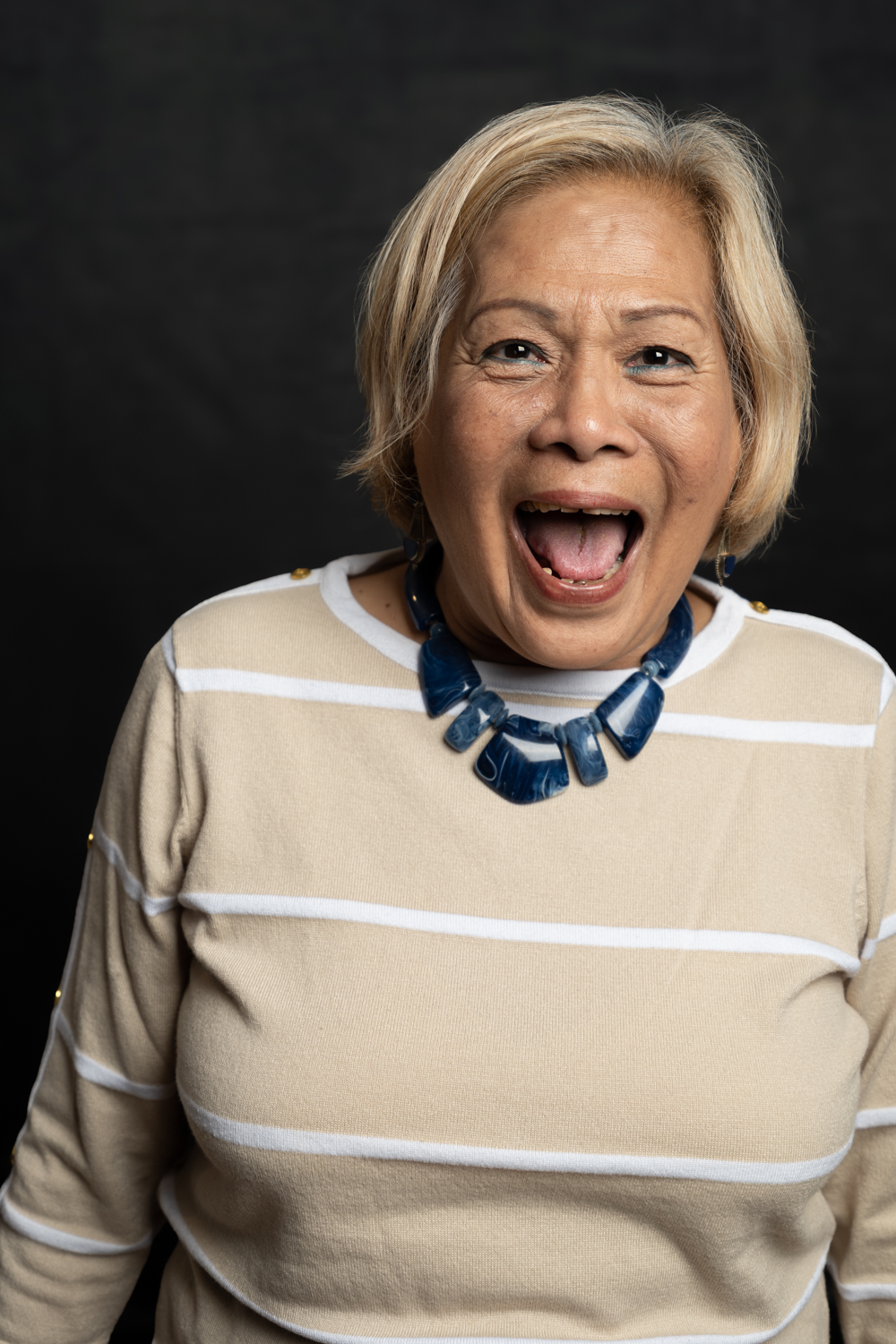 A photographic portrait of a woman with a very wide smile and short blonde hair. She is wearing a beige and white striped top, with a large blue necklace.
