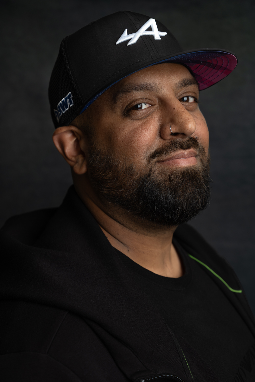 A photographic portrait of a man wearing a black LA baseball cap and a black hoodie. He is smiling, has a nose piercing and a short black beard.