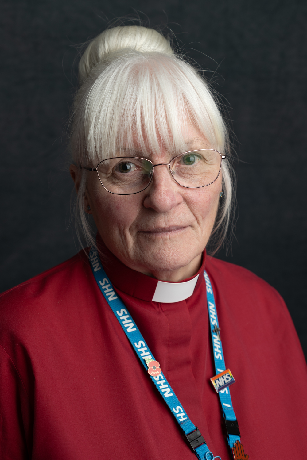 A photographic portrait of a woman wearing red scrubs, with a clerical collar and a blue NHS lanyard. Her white hair is in a bun with her fringe loose, and she is wearing glasses.