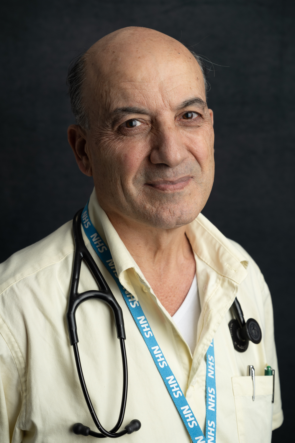 A photographic portrait of a bald man in a pale yellow shirt. He is smiling gently, wearing a blue NHS lanyard and black stethoscope around his neck.