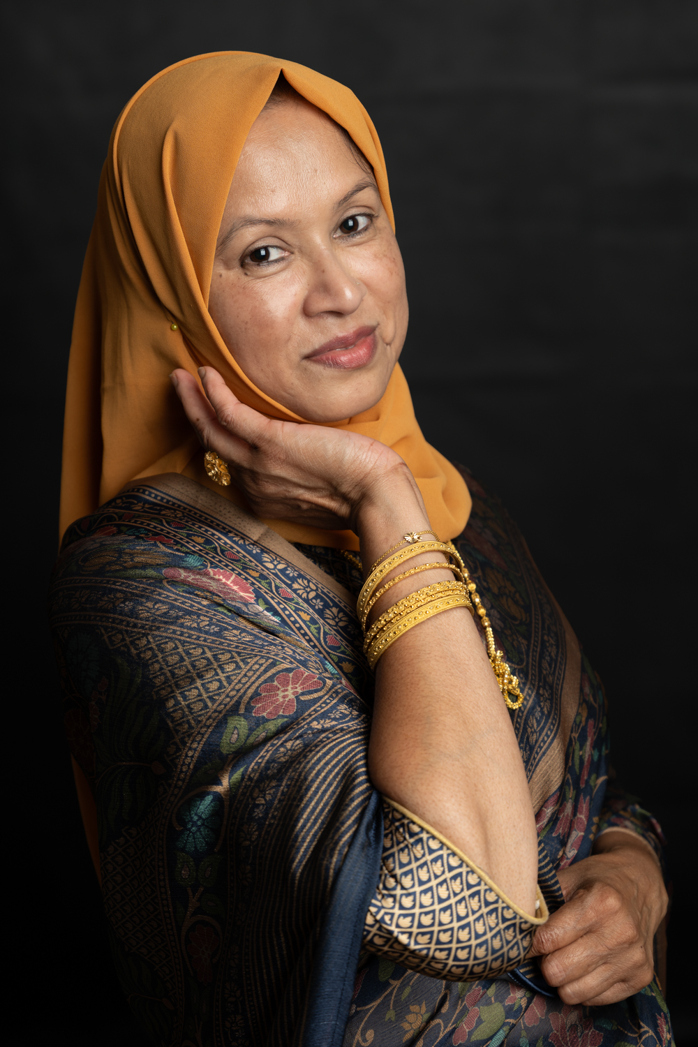 A photographic portrait of a woman in an orange hijab and blue patterned top. She has her hand to her cheek to show gold jewelry on her wrists and is smiling.