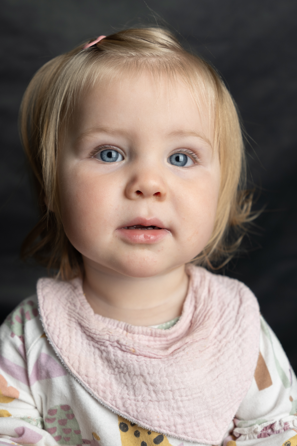 A photographic portrait of a toddler, with blonde hair and large blue eyes. She is wearing a pink and white outfit, including a hair clip and bib.