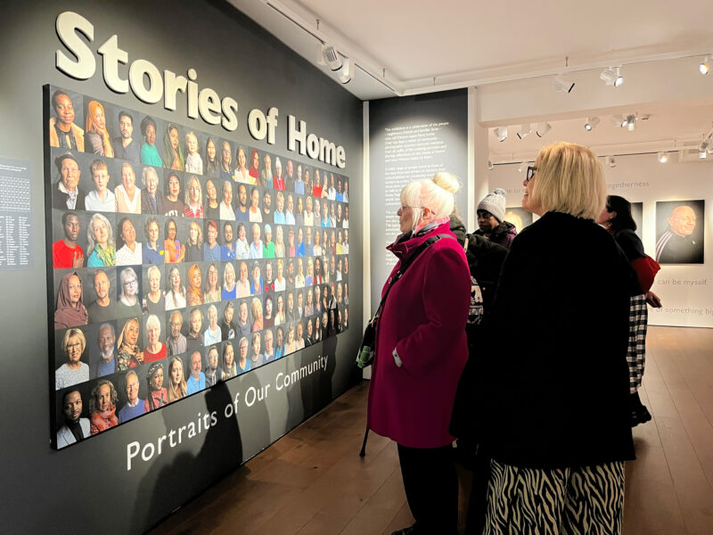 Two women in coats looking at a collage of photographic portraits which is mounted on the wall of a gallery.