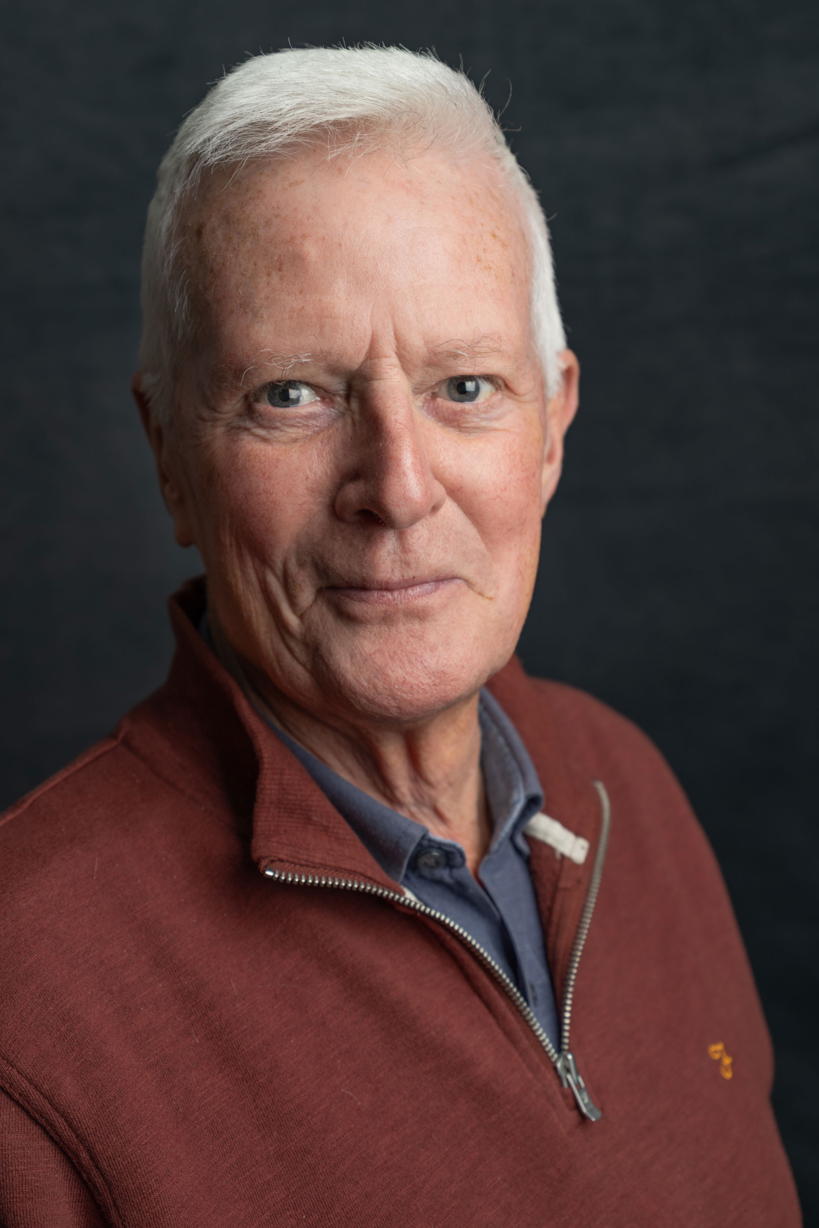 A photographic portrait of a man with short white hair, wearing a maroon quarter zip jumper and a blue polo top. He is smiling and looking directly into the camera.