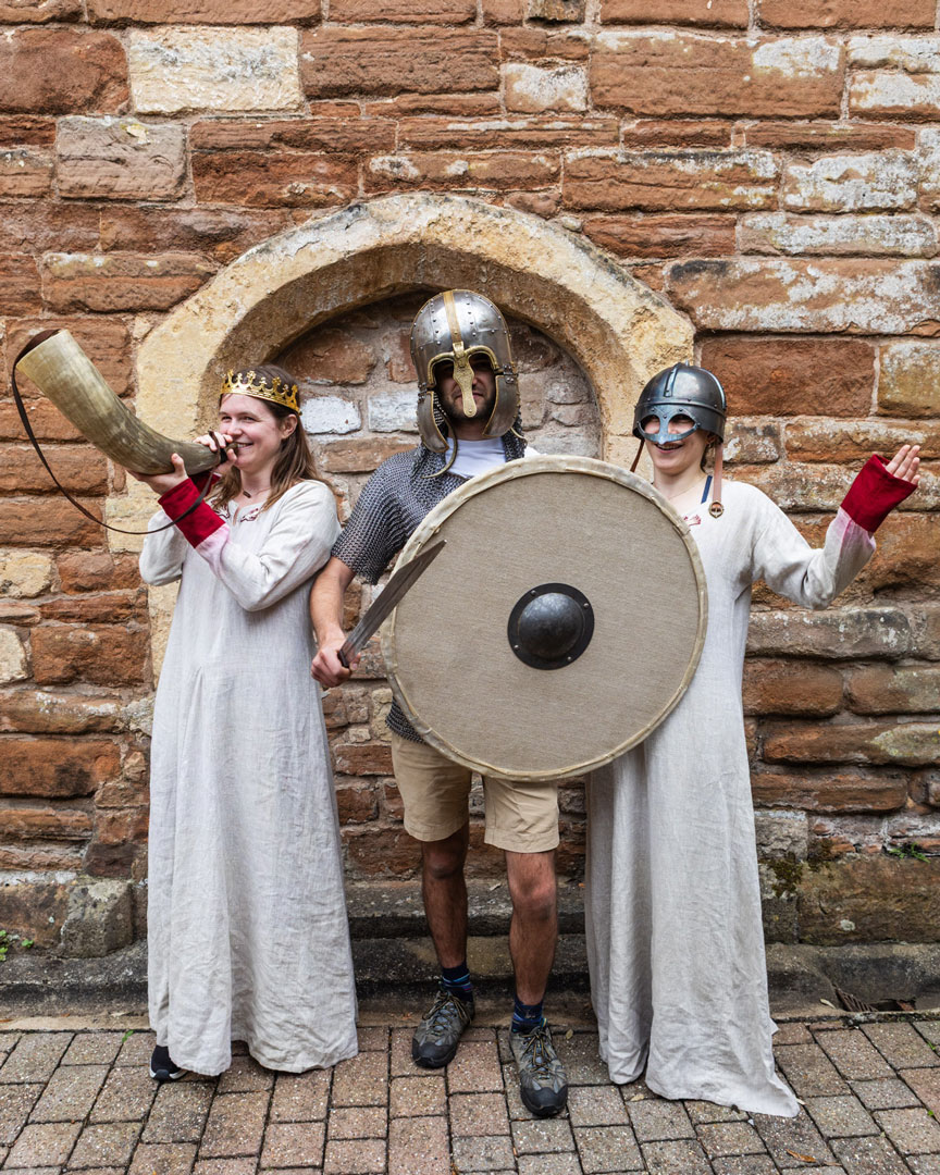 Three people dressed up in mediaval costume, wielding a horn and a shield.