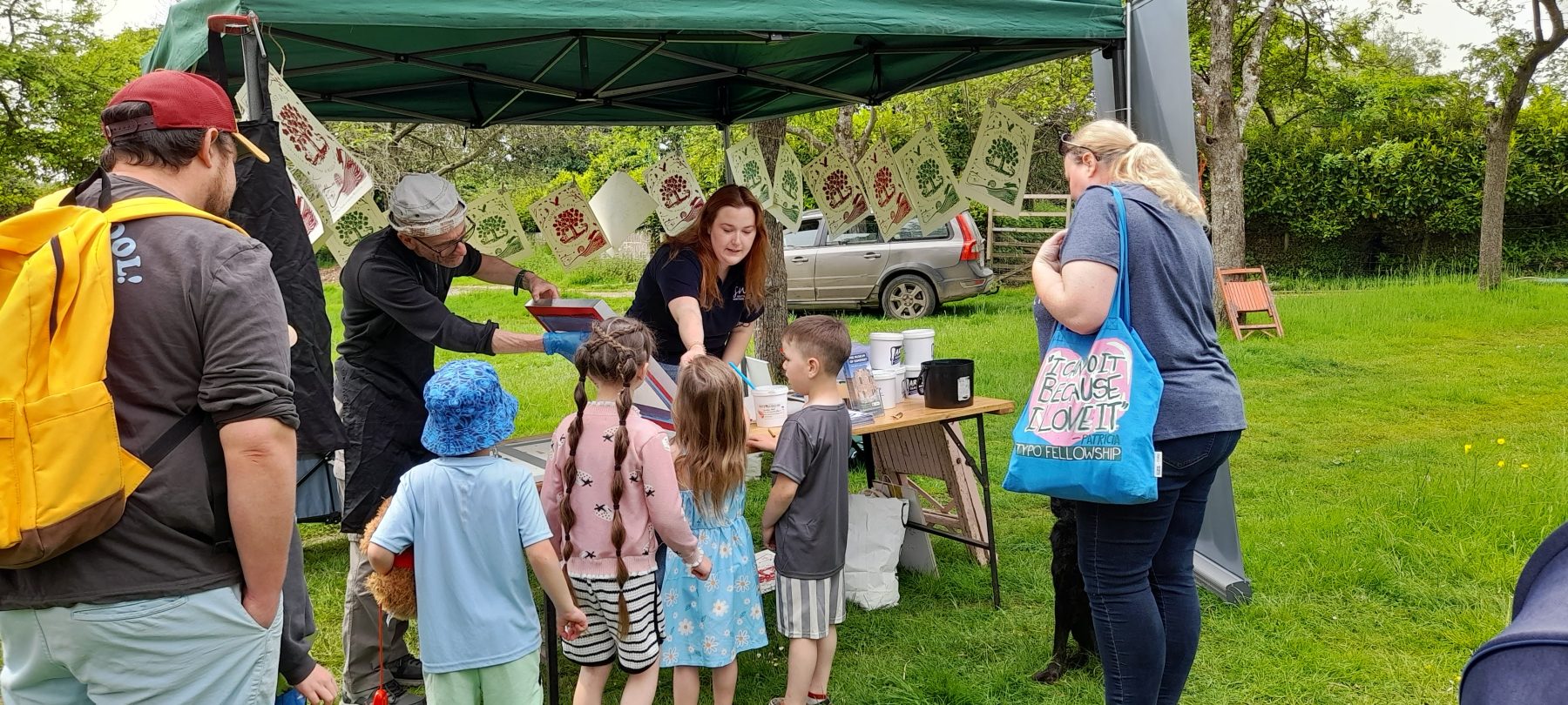 A group of two adults and four small children standing in front of a stall where two team members are demonstrating screen printing. They are outside in a green field, with prints hanging on a line behind them.