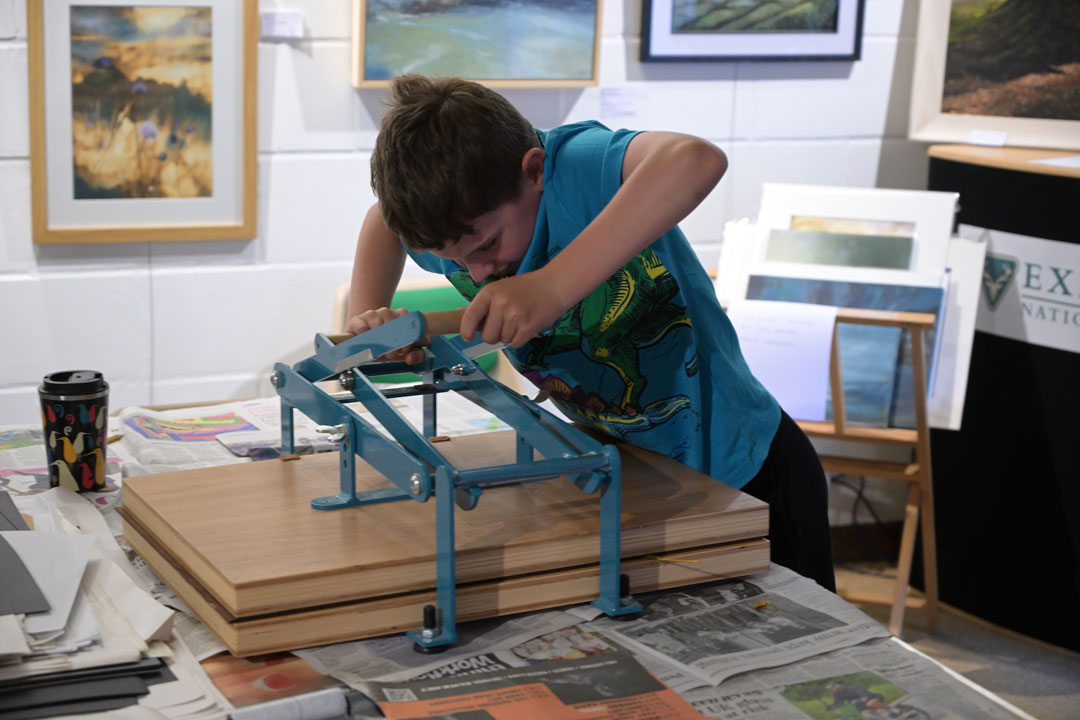 A young boy in a blue t-shirt leaning over a large printing press. There is newspaper on the table and artworks hung on a wall in the background.