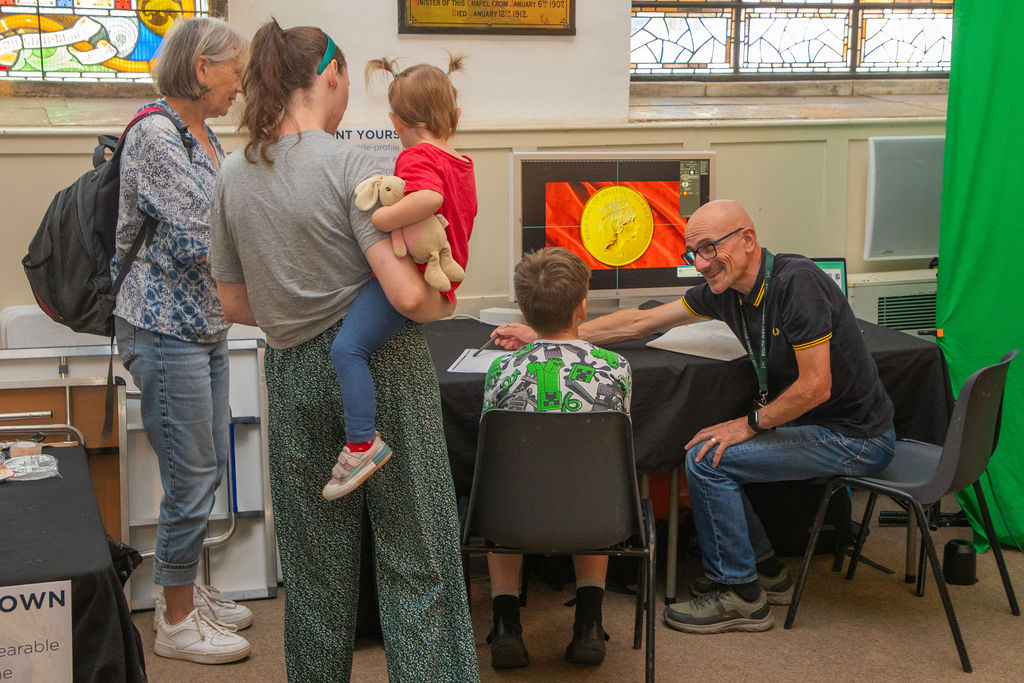 A group of adults and children gathered around a computer showing an editied picture of a young boy on a coin.