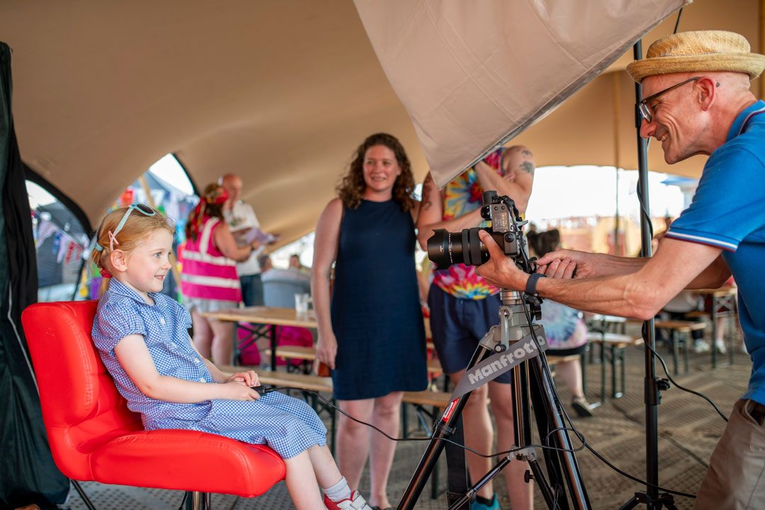 A little girl taking part in a self-portrait activity. She is sat in front of the camera on a red chair, with her parents and the activity facilitator watching on.